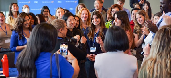 A large, diverse group of people stands in a circle, smiling, cheering, and clapping around a central interaction. Several individuals wear conference badges, and the atmosphere is lively and celebratory.