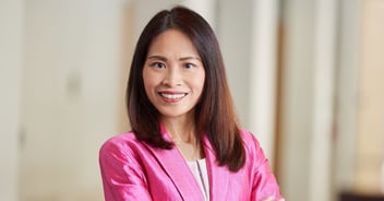 Christie K. Chung smiles at the camera. She is wearing a bright pink blazer over a light top, standing with her arms crossed in a softly lit indoor space.