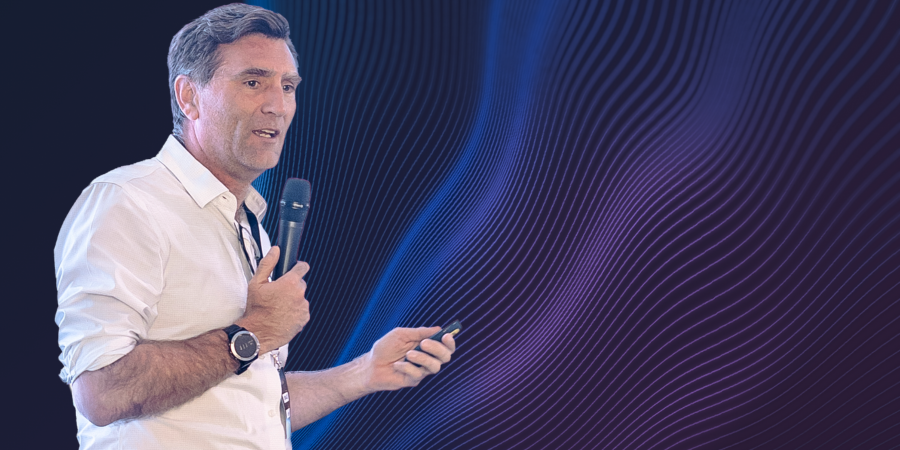 Speaker presenting while holding a microphone and presentation remote, standing against an abstract blue and purple wave graphic background.
