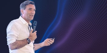 Speaker presenting while holding a microphone and presentation remote, standing against an abstract blue and purple wave graphic background.