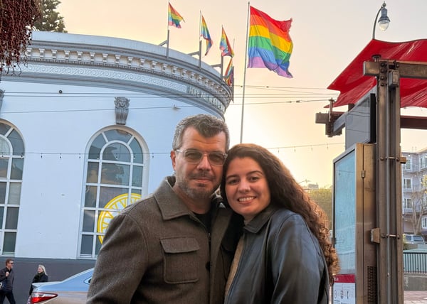 Marcos Apolonio stands with his daughter in San Francisco’s Castro District, smiling at the camera with rainbow pride flags flying above the Harvey Milk Plaza building in the background, photographed in March 2025.