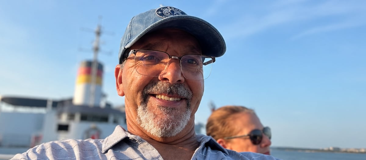 A smiling man takes a selfie while riding on a ferry under a clear blue sky.
