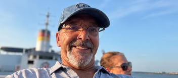 A smiling man takes a selfie while riding on a ferry under a clear blue sky.