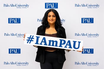 Shirin Aghakhani poses in front of a Palo Alto University backdrop holding an "#IAmPAU" sign.
